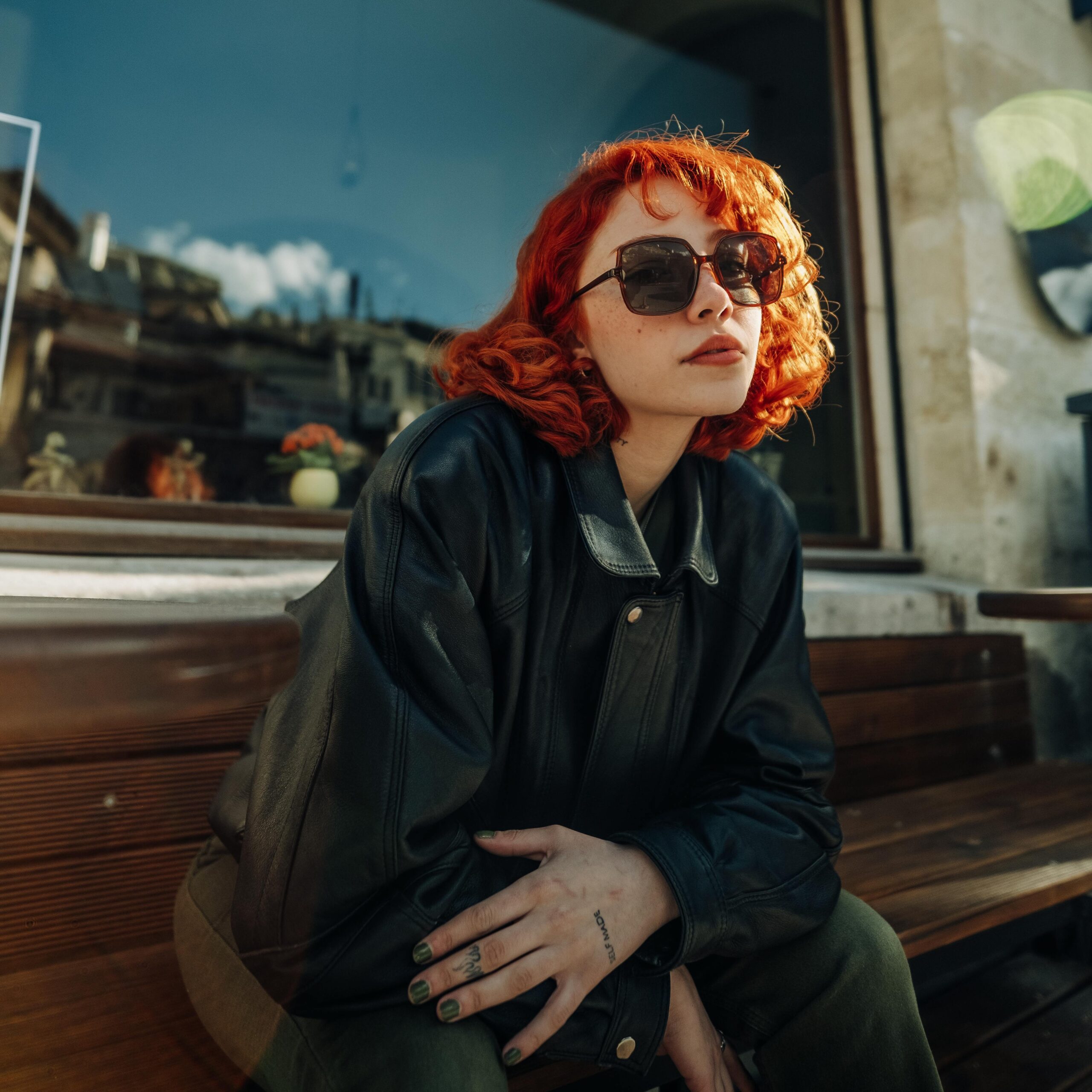 Young Redhead in a Leather Jacket and Sunglasses Sitting on a Bench